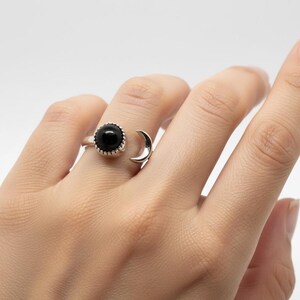 May include: A silver open-style ring featuring a round, polished black stone set in a scalloped bezel on one end and a smooth, crescent moon shape on the other end. The band is crafted from a polished silver-toned metal with a sleek finish.