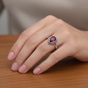 May include: A silver ring featuring a pear-shaped, deep magenta gemstone set in a bezel with a beaded border. The band is smooth and polished, showcasing a classic design that highlights the faceted stone and the intricate metalwork surrounding it.