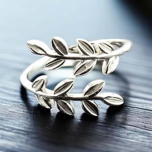 May include: A silver-colored ring featuring a leaf design. The ring wraps around the finger with two sets of leaves facing opposite directions. The leaves have detailed veining, and the ring is set against a dark background.