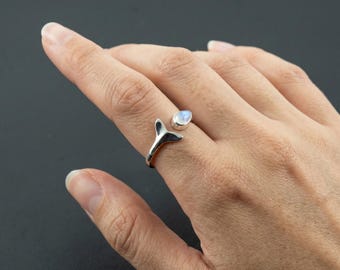 May include: A silver open-style ring featuring a polished whale tail motif on one end and a small, smooth, iridescent oval moonstone cabochon on the other end. The band is crafted from a reflective silver-toned metal with a smooth, curved finish.