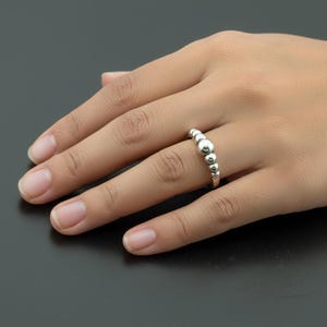May include: A silver-toned ring featuring a series of polished spherical beads of varying sizes arranged in a row along the band. The largest bead is positioned in the center, with smaller beads tapering off toward the sides of the metal ring.