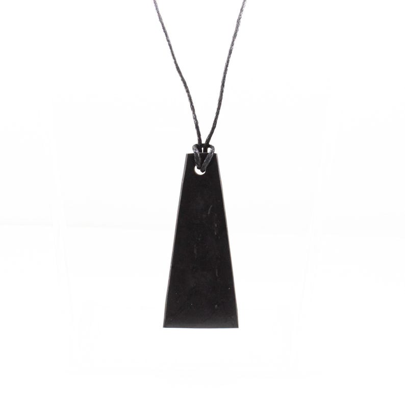 May include: A black trapezoidal pendant suspended from a thin black cord. The pendant has a smooth, matte finish and a single circular hole at the top center where the cord passes through, creating a simple and clean geometric necklace design.
