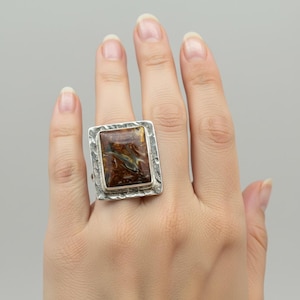 Puede incluir: Un anillo rectangular con una piedra pulida de tonos ámbar y marrón, engastada en un marco de plata martillada y texturizada. La piedra muestra patrones de remolinos en marrón oscuro, dorado y negro, sujeta por un borde metálico rústico.