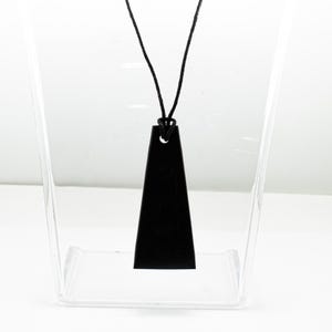 May include: A black pendant with a trapezoidal shape featuring a single hole at the top, suspended from a thin black cord. The pendant has a smooth, matte finish and a flat surface, hanging vertically from the center of the black cord loop.