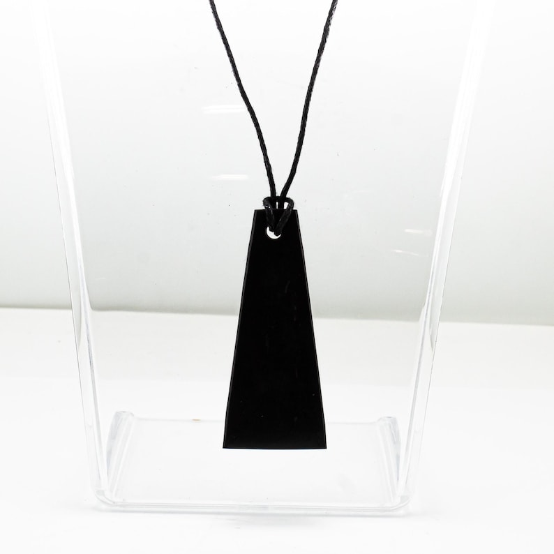May include: A black pendant with a trapezoidal shape featuring a single hole at the top, suspended from a thin black cord. The pendant has a smooth, matte finish and a flat surface, hanging vertically from the center of the black cord loop.