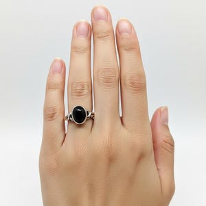 May include: A silver ring featuring a smooth, oval-shaped black stone set in the centre. The band of the ring has a decorative, twisted metal design that extends from the central setting, creating a detailed look around the finger.