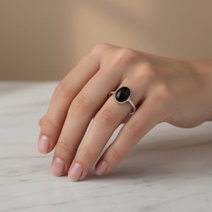 May include: A silver ring featuring a smooth, polished oval black stone set in a simple metal band. The ring is worn on the ring finger, showcasing the contrast between the dark, glossy gemstone and the metallic finish of the band.