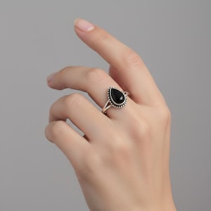 May include: A silver ring featuring a teardrop-shaped black stone set in a beaded metal frame. The band is split into two thin silver wires that meet at the base of the setting, creating a delicate and structured appearance on the finger.