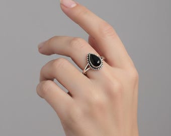 May include: A silver ring featuring a teardrop-shaped black stone set in a beaded metal frame. The band is split into two thin silver wires that meet at the base of the setting, creating a delicate and structured appearance on the finger.