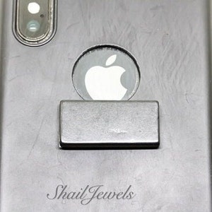 May include: A silver metal phone ring holder attached to the back of a black smartphone. The phone ring holder is rectangular in shape and has a smooth surface.