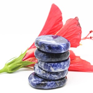 Pode incluir: Uma pilha de cinco pedras de sodalita azul lisas e polidas, cada uma com um padr&atilde;o veteado &uacute;nico. As pedras est&atilde;o dispostas em forma de pir&acirc;mide, com a pedra maior na parte inferior e a menor na parte superior. As pedras repousam sobre uma superf&iacute;cie branca, com uma flor de hibisco vermelha atr&aacute;s delas.