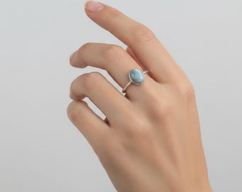 May include: A silver ring featuring a smooth, oval-shaped light blue stone set in a simple metal band. The stone has a marbled appearance with soft white and pale blue tones, securely held in a polished silver setting on a thin, rounded band.