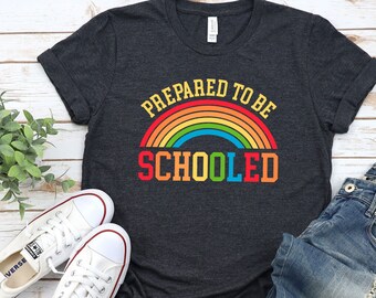 May include: A dark grey t-shirt with a rainbow graphic and the text "Prepared to be Schooled". The shirt is shown on a white wooden background with a pair of white trainers and blue jeans.