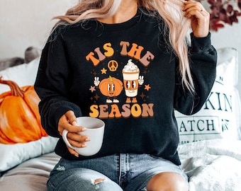May include: Black sweatshirt with a retro graphic reading "Tis the Season." The design includes a cartoon pumpkin, coffee cup, peace signs, and stars in orange, brown, and white. This is a cosy autumn-themed item of clothing.