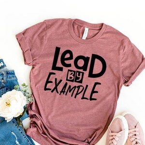 May include: A mauve t-shirt with the text "Lead by Example" in black lettering. The shirt is paired with blue jeans and pink sneakers.