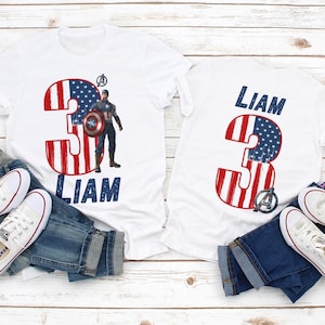 Peut inclure: T-shirts blancs avec un motif patriotique repr&eacute;sentant le chiffre 3 en rouge et bleu, avec le motif du drapeau am&eacute;ricain. Un t-shirt pr&eacute;sente un graphisme Captain America et le nom "Liam". Les t-shirts sont associ&eacute;s &agrave; des jeans bleus et des baskets blanches.