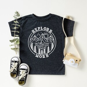 May include: A dark grey t-shirt with a white graphic of a mountain range, lake, sun, and trees in a circle with the text "Explore More".
