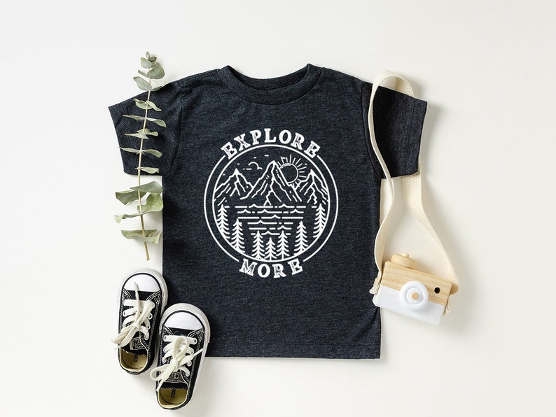 May include: A dark grey t-shirt with a white graphic of a mountain range, lake, sun, and trees in a circle with the text "Explore More".