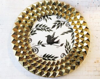 May include: A round, gold-colored frame with a geometric, studded design. The frame surrounds a black and white illustration of a bird perched on a branch with leaves. The artwork is centered on a white background.