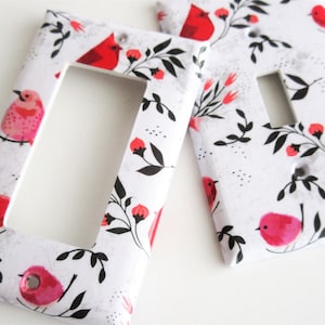 May include: Two white light switch covers featuring a red cardinal and pink bird design. The background is white with black floral accents.