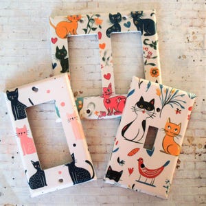 Op de afbeelding: Vier lichtschakelaars met een katten- en bloemenpatroon. De afdekkingen zijn wit met een roze, oranje en zwart kattenontwerp. De katten zijn omringd door bloemen en bladeren. De schakelaars zijn een leuke en eigenzinnige manier om een ​​persoonlijke touch aan je huis toe te voegen.