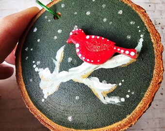 Könnte beinhalten: Ein runder Holzschmuck, der grün mit weißem Schnee bemalt ist und einen roten Vogel mit weißen Punkten auf einem Ast. Der Schmuck hat oben ein Loch zum Aufhängen.