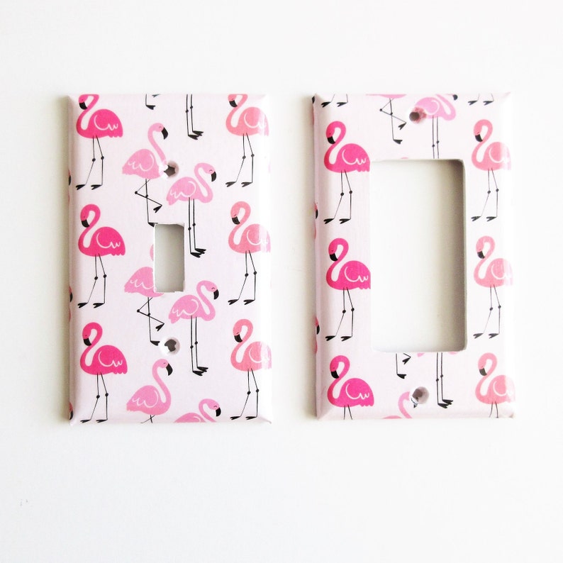 May include: Two decorative light switch covers featuring a pink flamingo pattern on a white backdrop. The flamingos are in different poses and shades of pink. One cover has a single switch opening, the other has a larger opening.