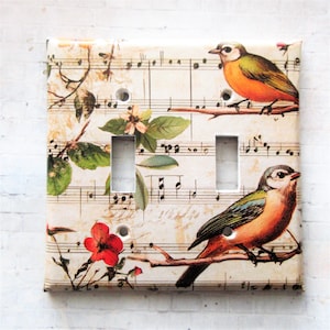 May include: A double light switch cover with a vintage design. The cover has a cream background with musical notes and illustrations of birds, flowers, and branches. The birds have orange, green, and blue feathers.