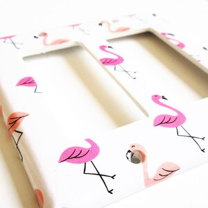 May include: A white double light switch cover with a repeating pattern of pink flamingos. The flamingos have black legs and beaks, with pink accents. The cover has two rectangular cutouts.