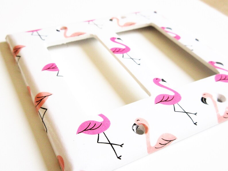May include: A white double light switch cover with a repeating pattern of pink flamingos. The flamingos have black legs and beaks, with pink accents. The cover has two rectangular cutouts.