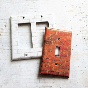 May include: Two light switch covers, one with a white brick pattern and one with a brown brick pattern. The brown brick switch cover has a single toggle switch.