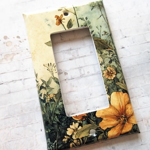 Könnte beinhalten: Dekorative Lichtschalterabdeckung mit floralem Design. Sie zeigt gelbe und orangefarbene Blumen, grüne Blätter und einen cremefarbenen Hintergrund. Die rechteckige Öffnung ist für einen Standardschalter vorgesehen. Maße in Zentimetern.