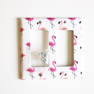 May include: A white double light switch cover with a repeating pattern of pink flamingos. The cover has two rectangular cutouts and a small packet of screws. The flamingos have black legs and beaks.