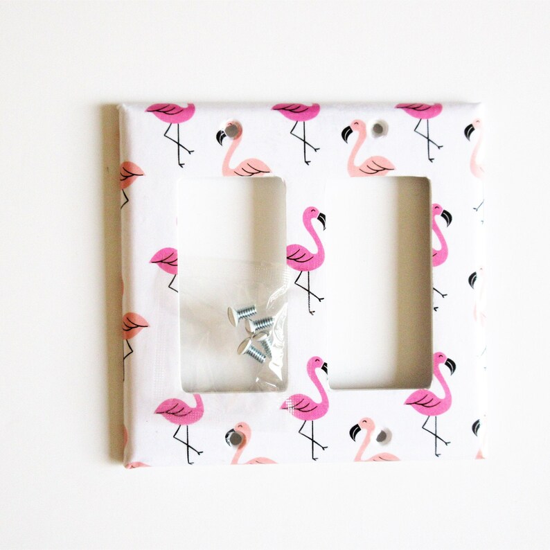 May include: A white double light switch cover with a repeating pattern of pink flamingos. The cover has two rectangular cutouts and a small packet of screws. The flamingos have black legs and beaks.