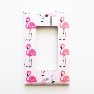 May include: A light pink single light switch cover with a repeating pattern of pink flamingos. The flamingos have black legs and beaks. The cover has a rectangular opening for the switch and a screw hole.