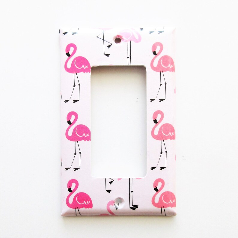 May include: A light pink single light switch cover with a repeating pattern of pink flamingos. The flamingos have black legs and beaks. The cover has a rectangular opening for the switch and a screw hole.