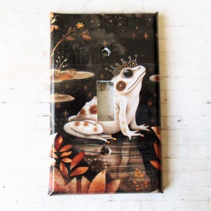 Puede incluir: Una tapa de interruptor de luz con una rana blanca con manchas marrones que lleva una corona dorada. La rana está sentada en un nenúfar en un estanque azul oscuro con estrellas doradas y un reflejo de la rana en el agua. El fondo es un bosque oscuro con hojas marrones y naranjas.