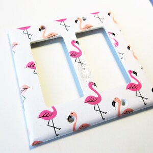 May include: White double light switch cover decorated with pink flamingo illustrations. The flamingos have black beaks and legs. The background is white.