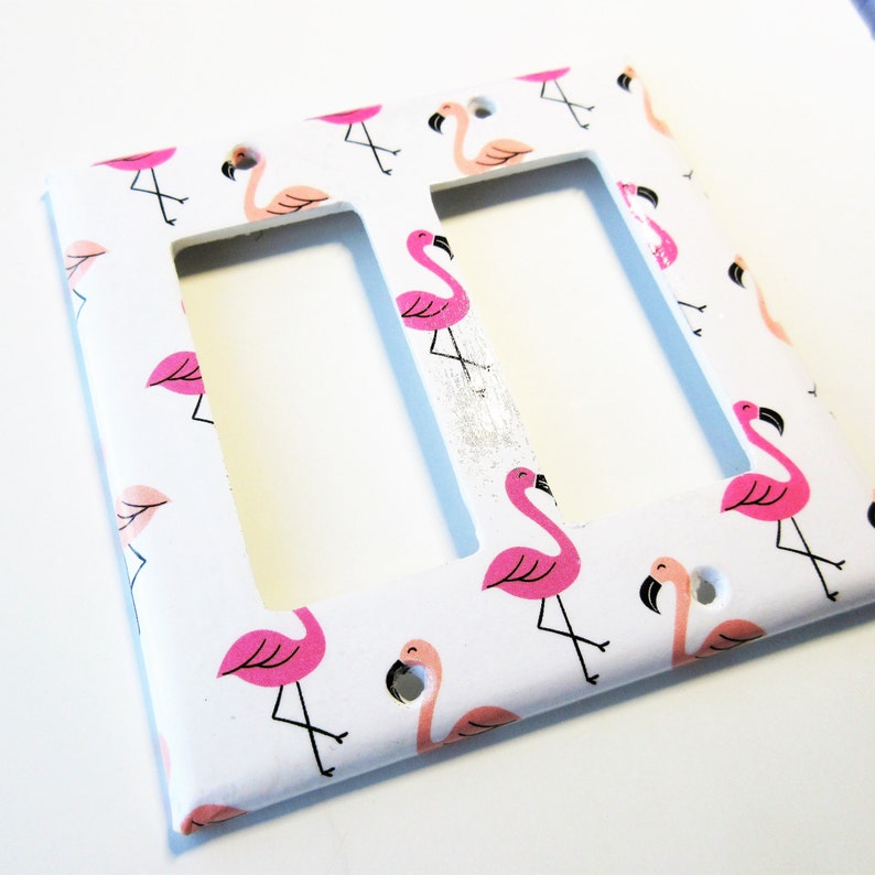 May include: White double light switch cover decorated with pink flamingo illustrations. The flamingos have black beaks and legs. The background is white.