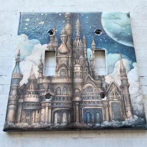 May include: A double light switch cover featuring a whimsical illustration of a fairytale castle with a blue sky, clouds, and stars. The castle is detailed with towers, windows, and a moon in the background.