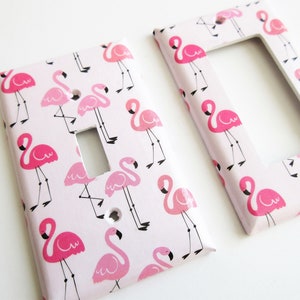 May include: Two decorative light switch covers featuring a pink flamingo pattern on a pale pink backdrop. The flamingos are depicted in various poses with black legs and beaks. One cover is rectangular, the other has a rectangular aperture.