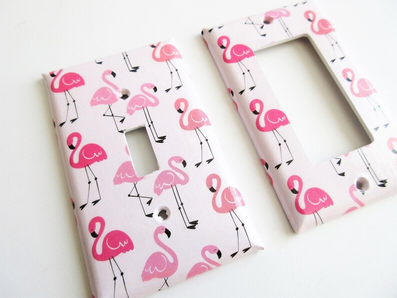 May include: Two decorative light switch covers featuring a pink flamingo pattern on a pale pink backdrop. The flamingos are depicted in various poses with black legs and beaks. One cover is rectangular, the other has a rectangular aperture.
