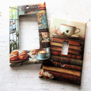 May include: Two light switch covers with vintage-inspired designs. One features a stack of old books with a cup of coffee and steam rising above it. The other features a cafe scene with pastries and a cup of cappuccino.