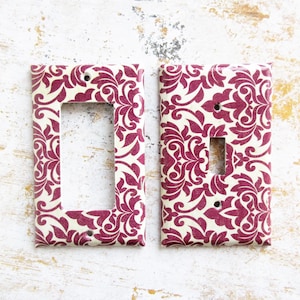 May include: Two decorative light switch covers with a white background and a burgundy damask pattern.
