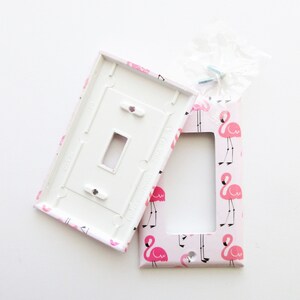 May include: A decorative light switch cover and matching plate with a pink flamingo pattern. The cover is white on the inside and has a rectangular opening. The flamingo design is repeated across the pink background.