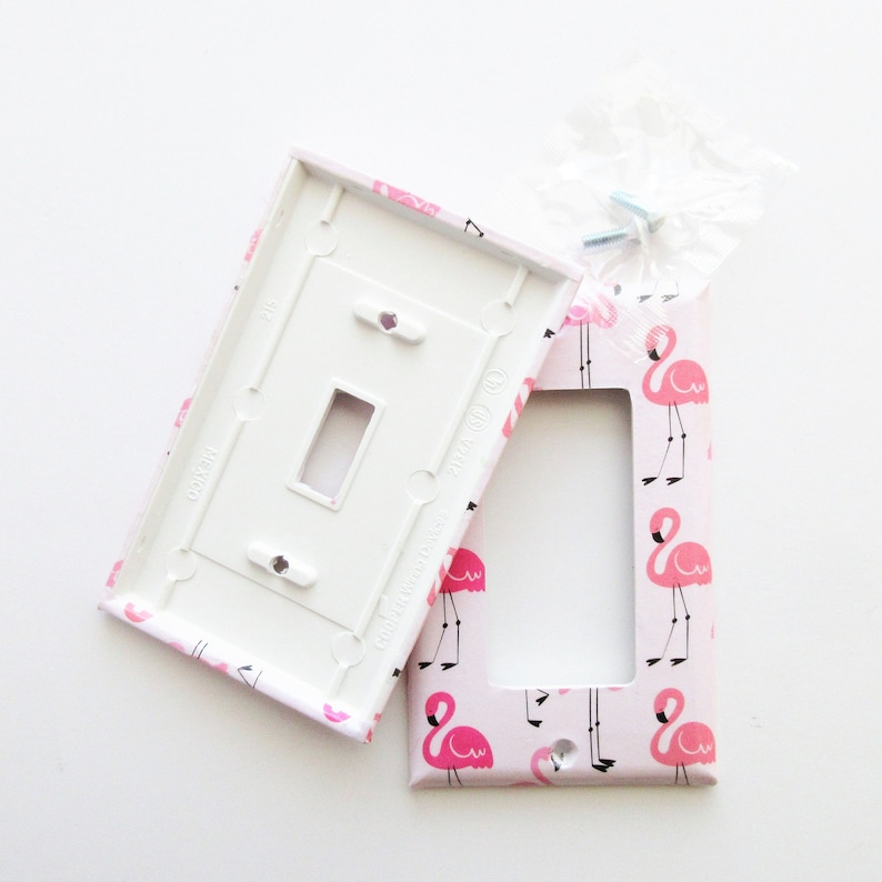 May include: A decorative light switch cover and matching plate with a pink flamingo pattern. The cover is white on the inside and has a rectangular opening. The flamingo design is repeated across the pink background.
