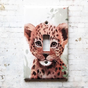 May include: A light switch cover featuring a watercolor painting of a brown and tan leopard cub with green foliage in the background.
