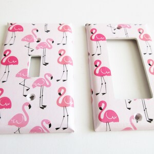 May include: Two light switch covers featuring a pink flamingo design on a pale pink backdrop. The flamingos are depicted in different poses, with black legs and beaks. One cover has a single switch aperture, the other has a double switch aperture.