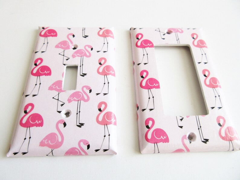May include: Two light switch covers featuring a pink flamingo design on a pale pink backdrop. The flamingos are depicted in different poses, with black legs and beaks. One cover has a single switch aperture, the other has a double switch aperture.