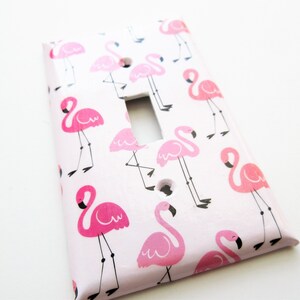 May include: A light pink switch plate decorated with a pattern of pink flamingos. The flamingos have black beaks and legs. The switch plate has a rectangular opening for the switch and two screw holes.